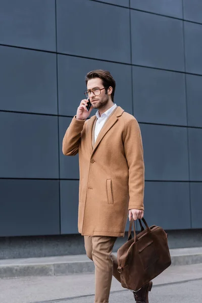 Young businessman in glasses walking with briefcase and talking on smartphone near building — Stock Photo