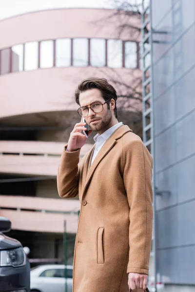 Young businessman in glasses talking on smartphone near building — Stock Photo