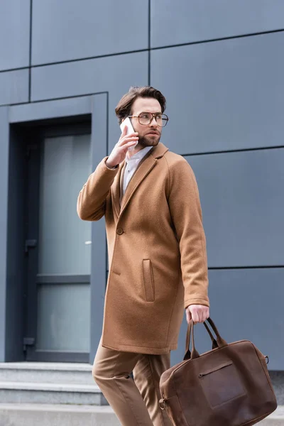 Young man in glasses and coat talking on smartphone near building — Stock Photo