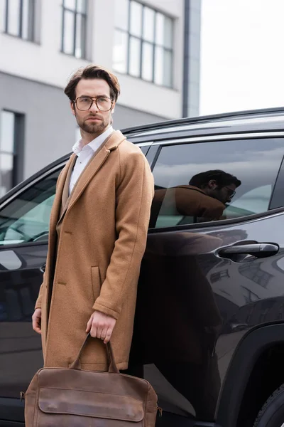 Young man in glasses and coat standing with briefcase near car — Stock Photo