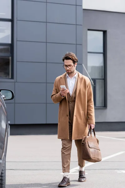 Junger Mann in Brille und Mantel steht mit Aktentasche und SMS auf Smartphone neben Auto — Stockfoto