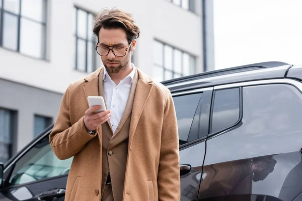 Hombre en gafas y el abrigo de mensajes de texto en el teléfono inteligente cerca del coche - foto de stock