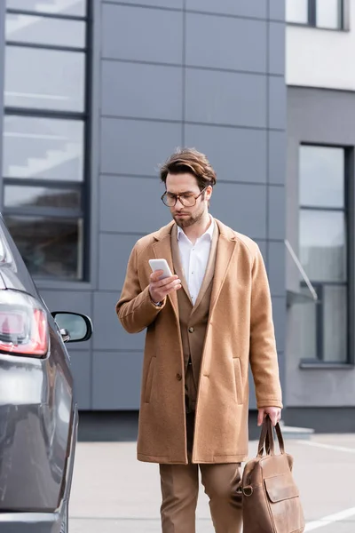 Businessman in glasses and coat texting on smartphone near car — Stock Photo