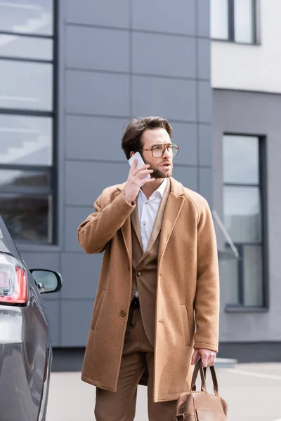 Hombre de negocios confiado en gafas y abrigo hablando por teléfono cerca del coche - foto de stock