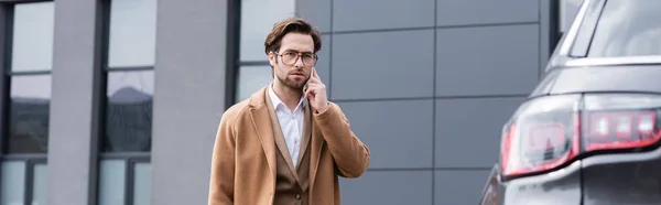 Hombre de negocios confiado en gafas y abrigo beige hablando por teléfono cerca del coche, pancarta - foto de stock
