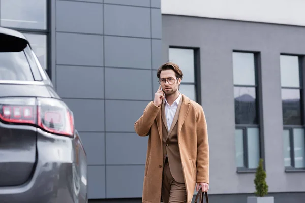 Confident businessman in glasses and beige coat talking on mobile phone near car — Stock Photo