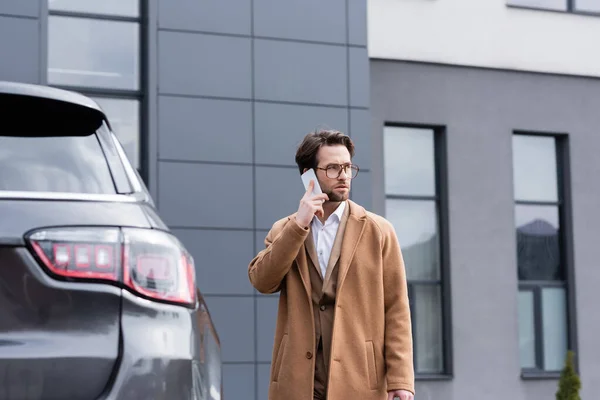 Уверенный в себе мужчина в очках и бежевом пальто разговаривает по мобильному телефону возле машины — стоковое фото