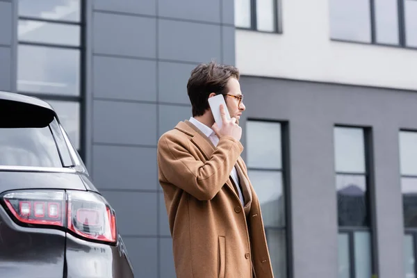 Seitenansicht eines selbstbewussten Mannes mit Brille und beigem Mantel, der in der Nähe des Autos mit dem Handy telefoniert — Stockfoto