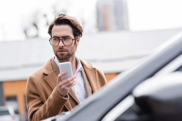 Confident man in glasses and beige coat holding mobile phone near blurred car — Stock Photo