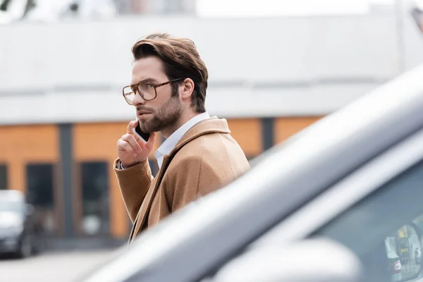 Young man in glasses and beige coat talking on mobile phone near blurred car — Stock Photo