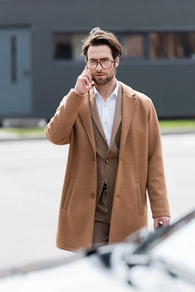 Уверенный в себе человек в очках и пальто разговаривает по мобильному телефону на открытом воздухе — стоковое фото