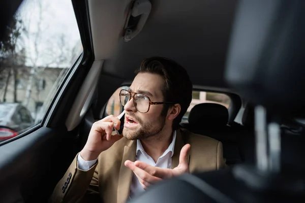 Hombre de negocios enojado en traje hablando en teléfono inteligente y mirando a la ventana del coche - foto de stock