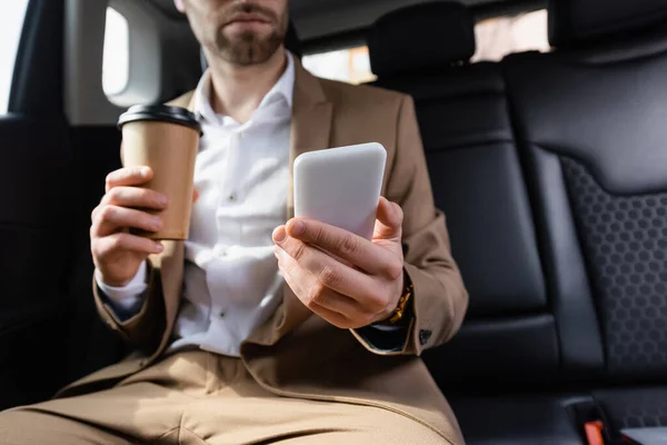 Vue recadrée de l'homme en costume tenant tasse en papier et smartphone en voiture — Photo de stock
