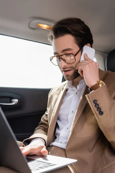 Empresário trabalhando remotamente com laptop enquanto conversa no smartphone no carro — Fotografia de Stock