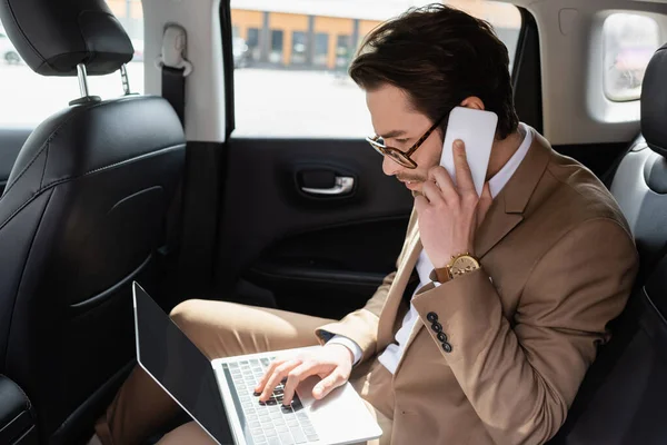 Uomo d'affari in occhiali che lavora in remoto con il computer portatile mentre parla su smartphone in auto — Foto stock