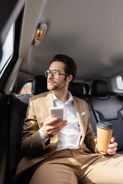 Hombre de negocios pensativo en traje y gafas con taza de papel y teléfono inteligente en el coche - foto de stock