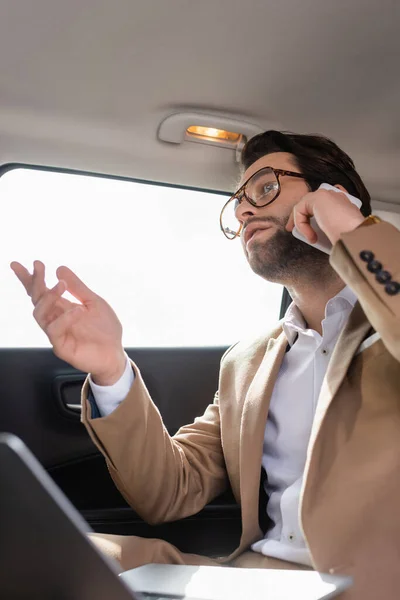 Vista angolo basso di uomo d'affari in occhiali parlare su smartphone vicino computer portatile offuscata in auto — Foto stock