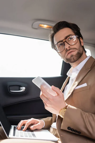 Uomo d'affari in occhiali che lavora in remoto con il computer portatile mentre tiene lo smartphone in auto — Foto stock