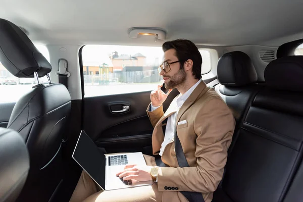 Hombre de negocios en gafas que trabajan a distancia con el ordenador portátil mientras habla en el teléfono celular en el coche - foto de stock