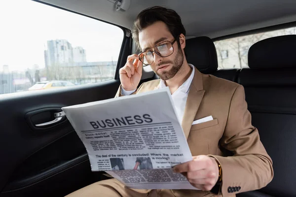 Uomo sicuro di sé che regola gli occhiali e legge il giornale aziendale in auto — Foto stock