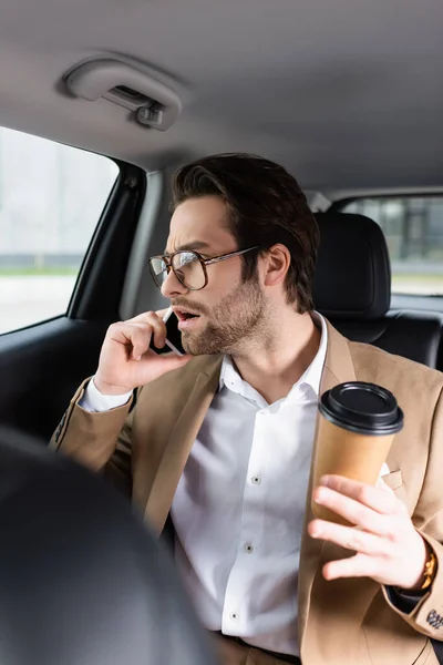 Hombre sorprendido en traje y gafas sosteniendo taza de papel y hablando en el teléfono inteligente en el coche - foto de stock