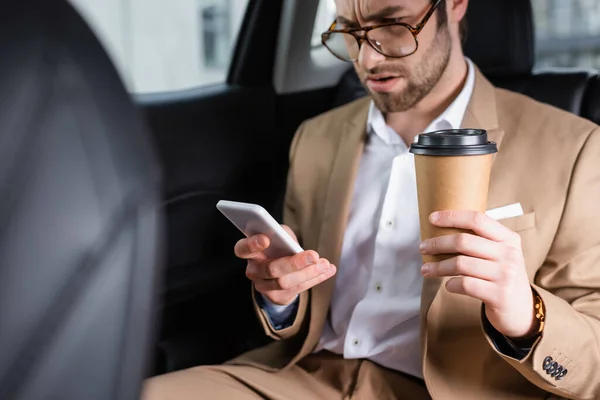Displeased man in suit and glasses holding paper cup and looking at smartphone in car — Stock Photo