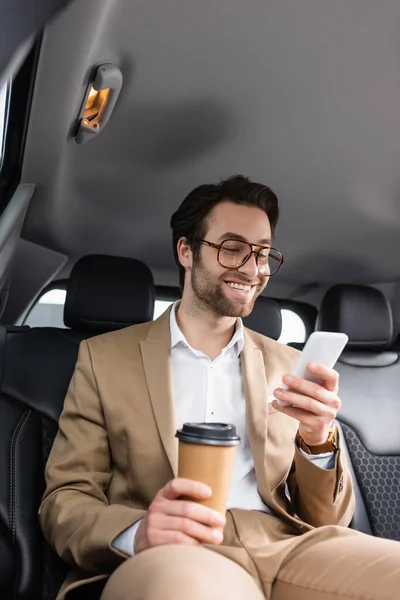Щасливий бізнесмен в костюмі і окулярах тримає паперовий стаканчик і смартфон в машині — стокове фото