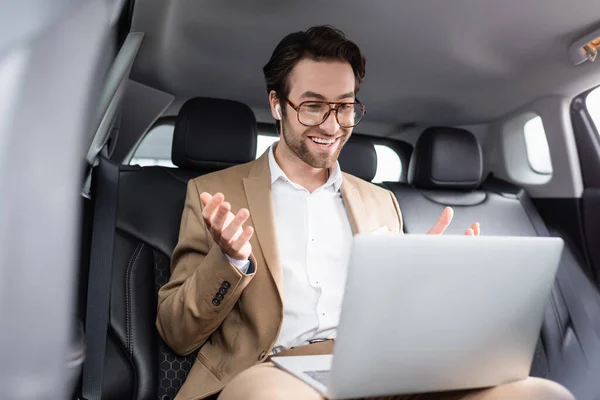 Hombre de negocios sonriente en gafas y auriculares inalámbricos gesticulando durante la videollamada en el coche - foto de stock