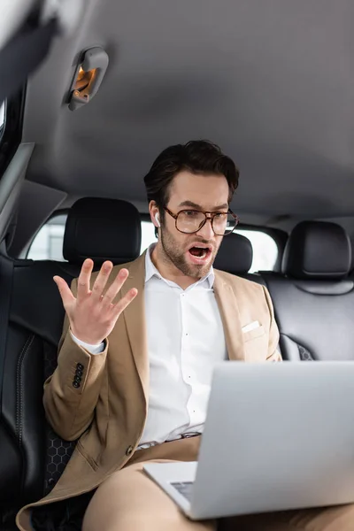 Homme d'affaires choqué dans des lunettes et écouteurs sans fil gestuelle lors d'un appel vidéo dans la voiture — Photo de stock