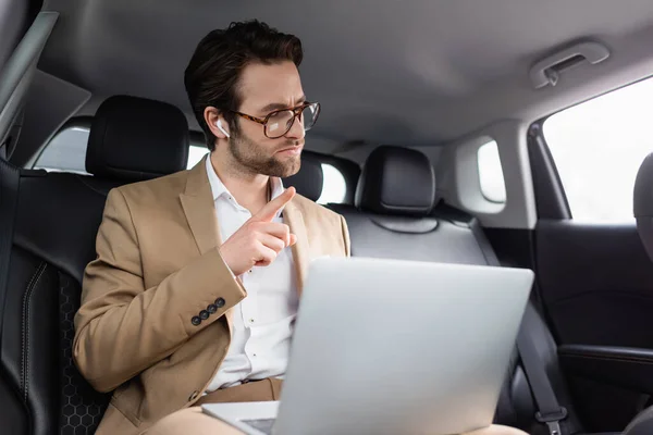 Geschäftsmann mit Brille und drahtlosen Kopfhörern zeigt bei Videoanruf im Auto mit dem Finger — Stockfoto