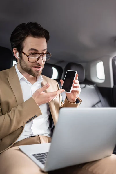 Businessman in glasses and wireless earphones pointing at smartphone during video call in car — Stock Photo