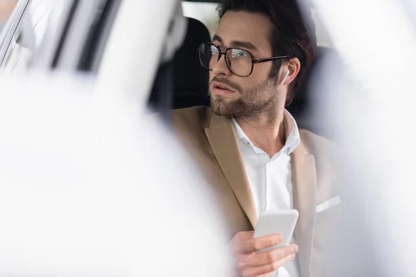 Homme d'affaires en lunettes et écouteurs sans fil tenant smartphone en voiture — Photo de stock