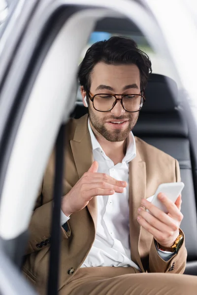 Businessman in glasses and wireless earphones gesturing during video call on smartphone inside of car — Stock Photo