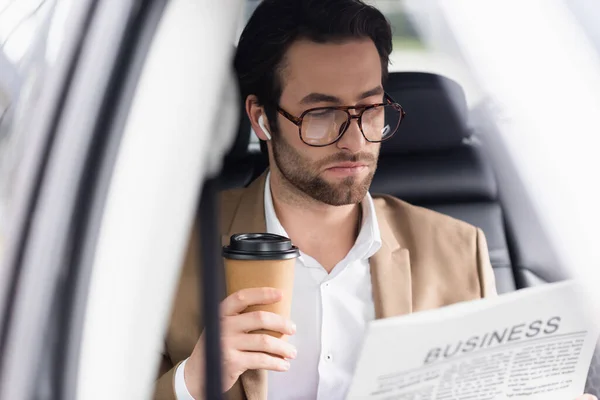 Bearded man in suit reading business newspaper and holding coffee to go in car — Stock Photo