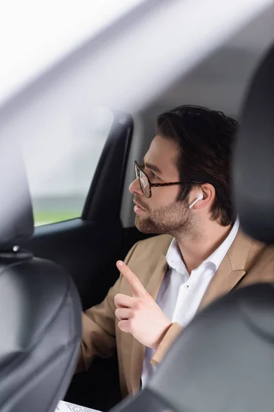 Uomo barbuto in occhiali e auricolari senza fili che punta con dito in auto — Foto stock