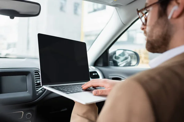 Hombre de negocios borroso en los auriculares inalámbricos usando el ordenador portátil con la pantalla en blanco en el coche - foto de stock