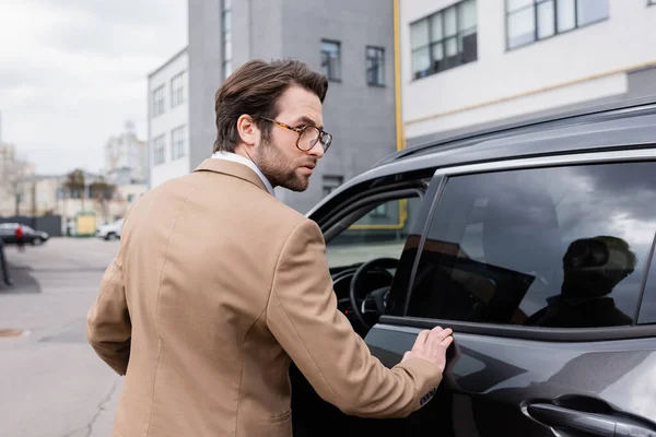 Hombre barbudo con chaqueta y gafas de pie cerca del coche con la puerta abierta — Stock Photo