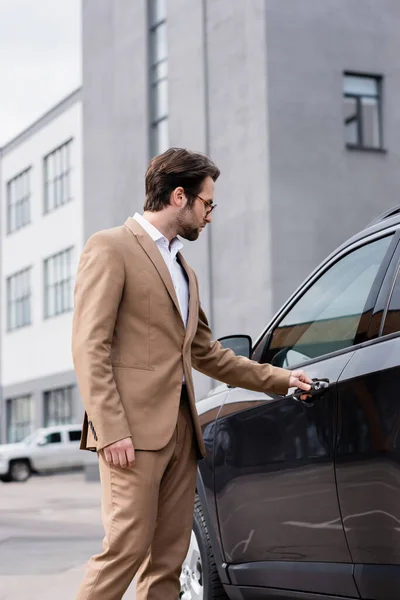 Homme barbu en veste beige et lunettes atteignant la porte de la voiture — Photo de stock