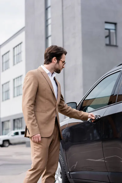 Hombre barbudo en chaqueta beige y gafas que llegan a la puerta del coche - foto de stock