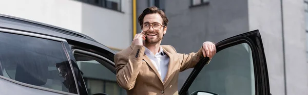 Hombre feliz con chaqueta y gafas hablando en el teléfono inteligente y de pie cerca del coche con la puerta abierta, pancarta - foto de stock