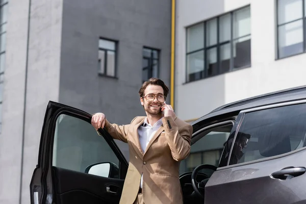 Веселий чоловік в костюмі і окулярах говорить на смартфоні і стоїть біля машини з відкритими дверима — стокове фото