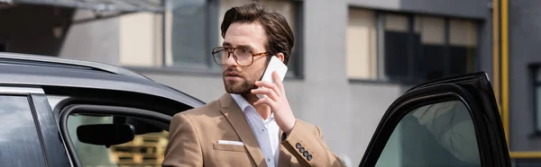 Бородатый мужчина в костюме и очках разговаривает по мобильному телефону и стоит рядом с машиной с открытой дверью, баннер — стоковое фото