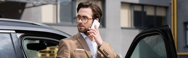 Hombre barbudo en traje y gafas hablando en el teléfono celular y de pie cerca del coche con la puerta abierta, pancarta — Stock Photo