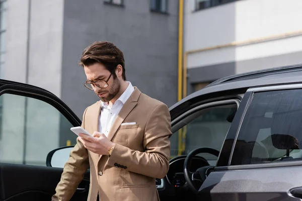 Homme barbu en costume et lunettes regardant le téléphone portable et debout près de la voiture avec porte ouverte — Photo de stock