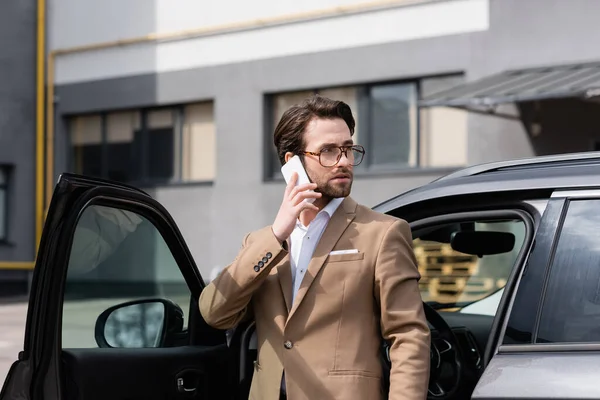 Hombre de negocios barbudo con traje y gafas hablando por teléfono celular y de pie cerca del coche con la puerta abierta - foto de stock