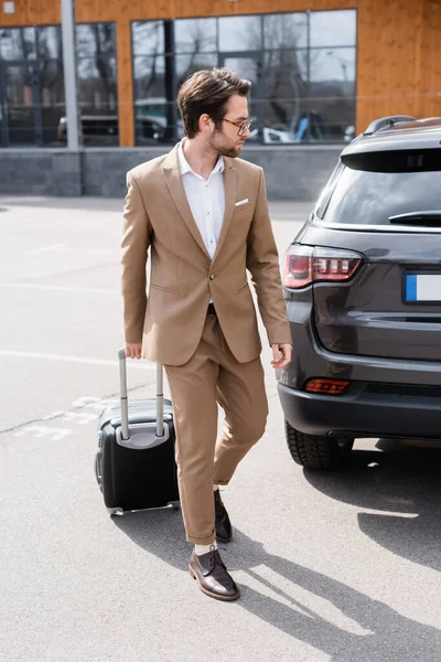 Full length of bearded man in suit and glasses walking with suitcase near car on parking — Stock Photo