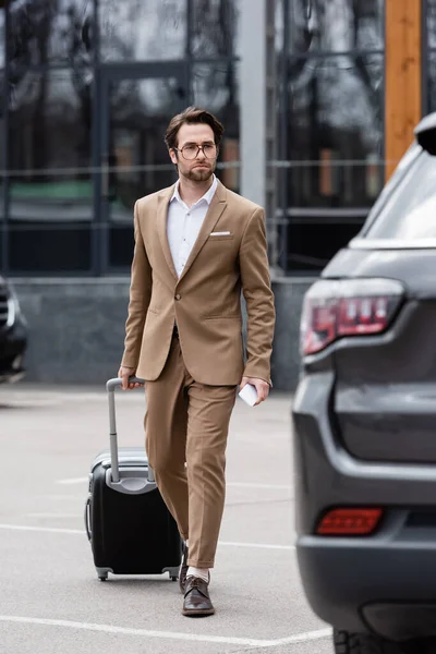 Full length of businessman in suit and glasses walking near car with suitcase — Stock Photo