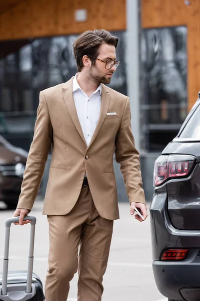 Homme en costume beige et lunettes tenant smartphone et debout près de la voiture avec valise — Photo de stock