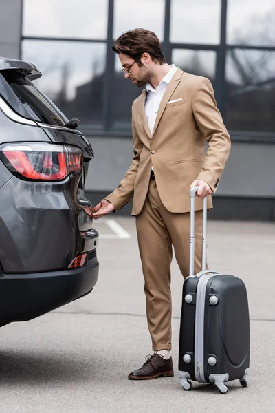 Чоловік у костюмі стоїть з валізою і відкриває автомобільний багажник — стокове фото