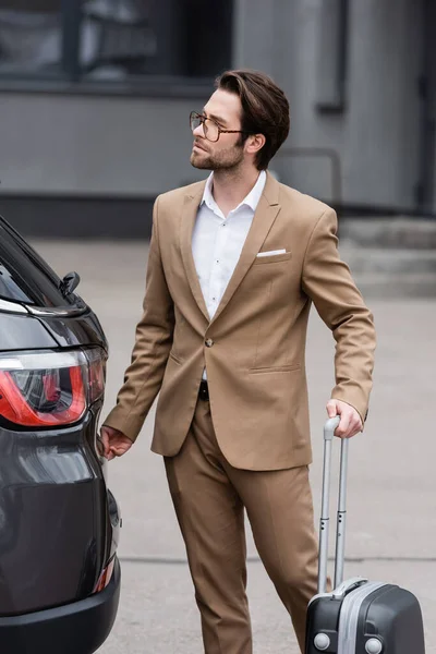 Uomo in giacca e cravatta in piedi con valigia vicino auto moderna — Foto stock